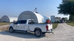 White pickup truck with “Anchor Roofing & Landscaping” signage parked near curved metal buildings in a rural area with grassy fields and clear skies in Omaha