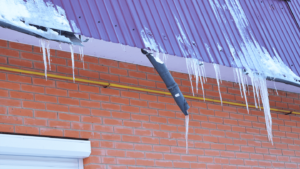 Icicles hanging from a snowy roof with a red corrugated sheet and a brick wall | Anchor Roofing | Omaha, Nebraska