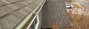 Overhead shot of a shingle roof with white gutters running along the edge. The roof is gray, and a portion of the house and a street are visible in the background.