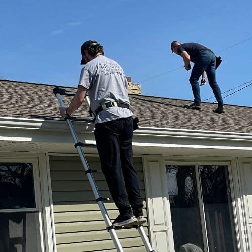 Two roof contractors on a roof, one on a ladder near the gutter and the other standing on the shingles in a process of roof replacement omaha.