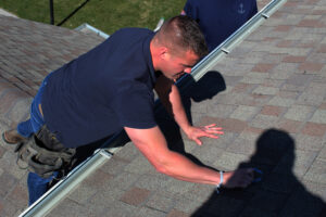 Worker kneeling on asphalt shingle roof, inspecting surface near the edge for possible damage in Omaha