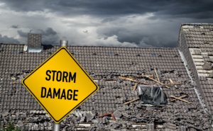 Residential roof with missing shingles and storm debris under dark clouds; yellow caution sign labeled ‘STORM DAMAGE’ placed on roof in Omaha