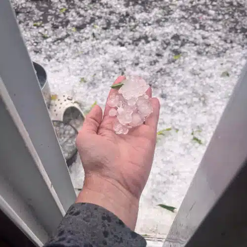 Hand holding large hailstones outside a Gretna, NE home after a recent storm, with yard covered in ice—demonstrating the intensity of hail that causes roofing hail damage in Omaha.