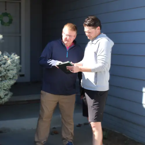 Two people standing outside a house reviewing information together on a tablet during daylight.
