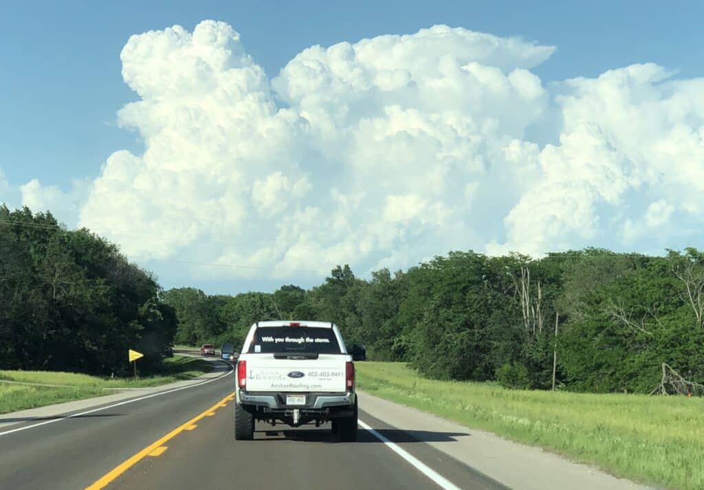 Branded service truck driving down a highway toward massive storm clouds to provide apartment roofing Omaha emergency roof repair in Omaha by Anchor Roofing