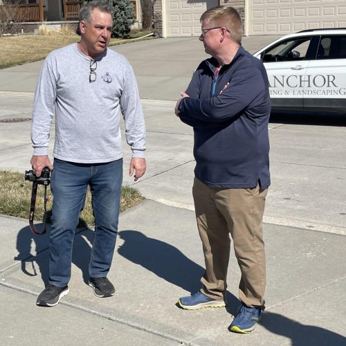 Two Anchor employees standing on a residential driveway discussing roof inspection; one holds a camera beside a vehicle marked ‘ANCHOR in Omaha